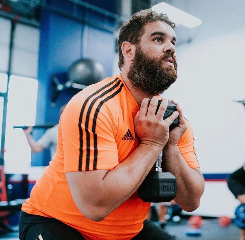 Man in orange athletic shirt holding a dumbbell at his chest in a gym