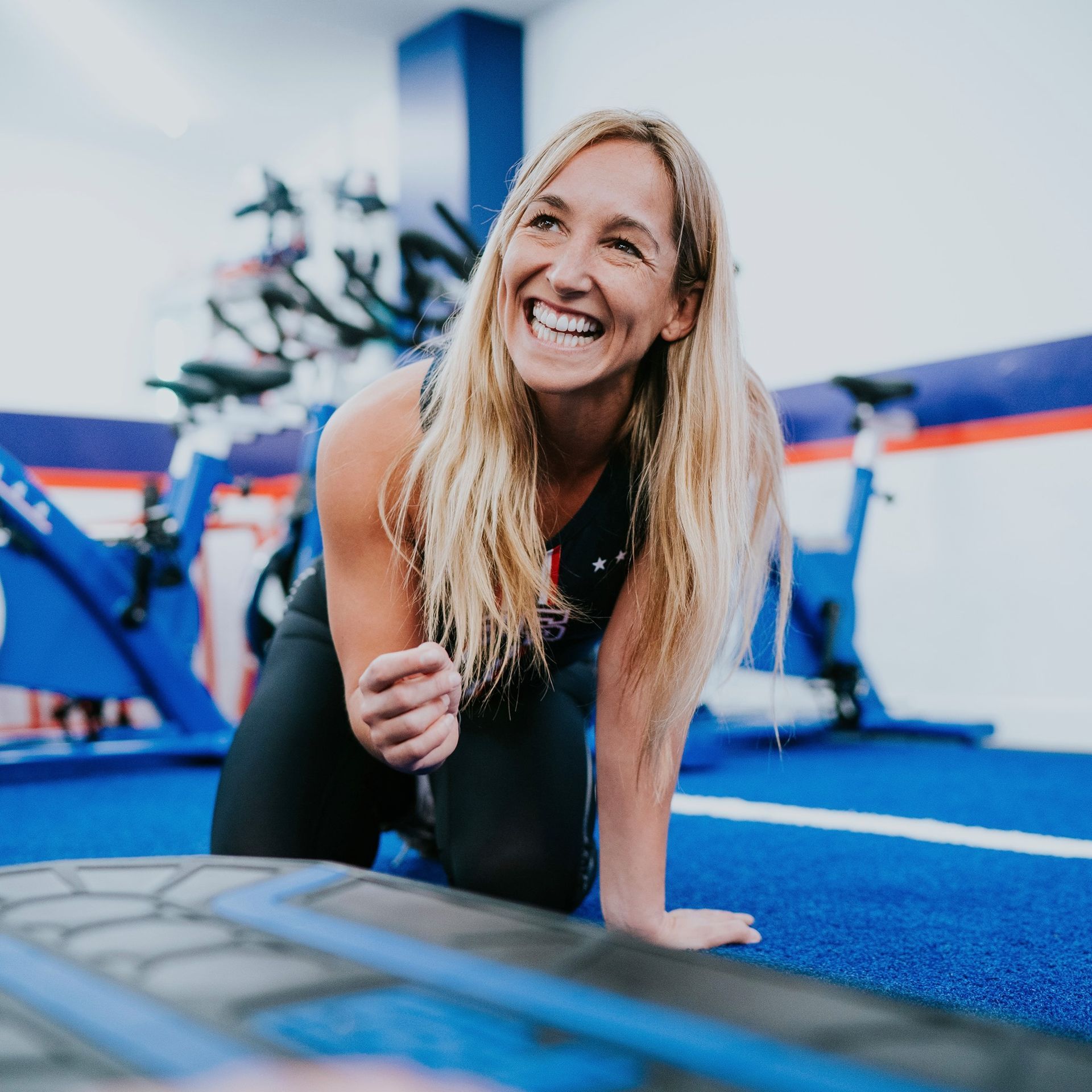 Smiling woman kneels on blue gym flooring in a fitness studio, leaning on one hand.
