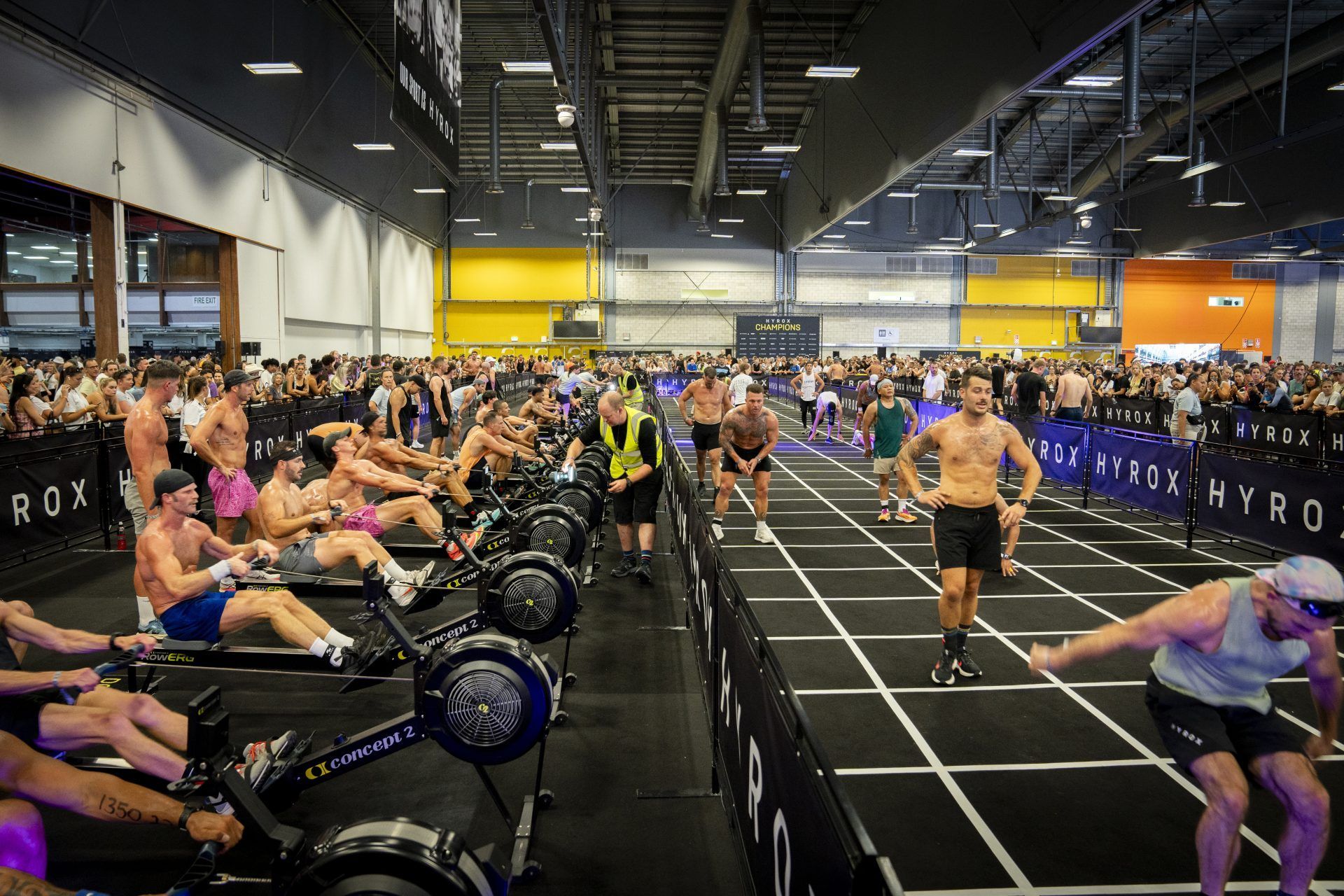 Large indoor rowing workout class with many people on rowing machines in a warehouse-style gym