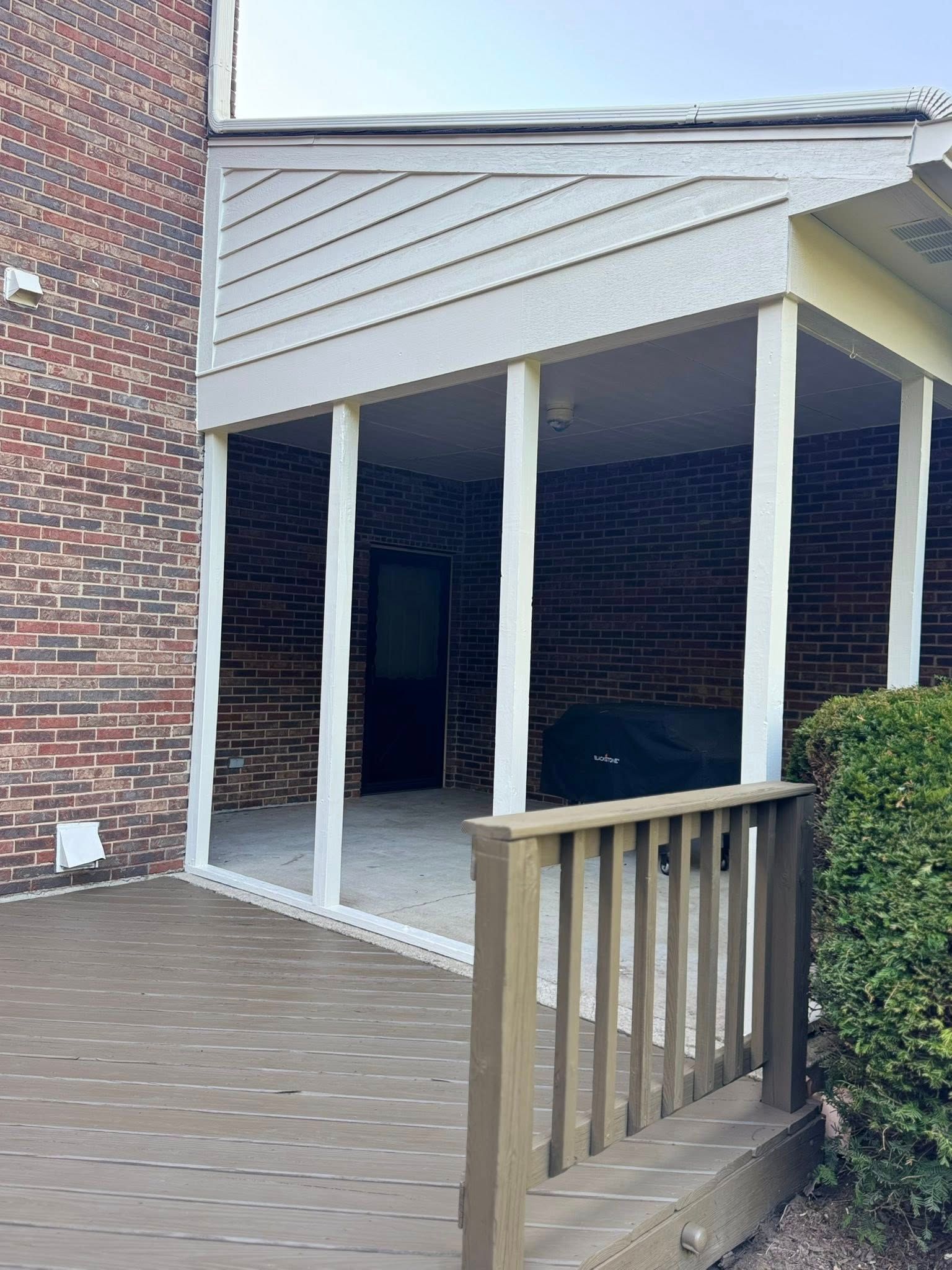 Covered outdoor patio with white pillars, adjacent to a brick wall and wooden deck.