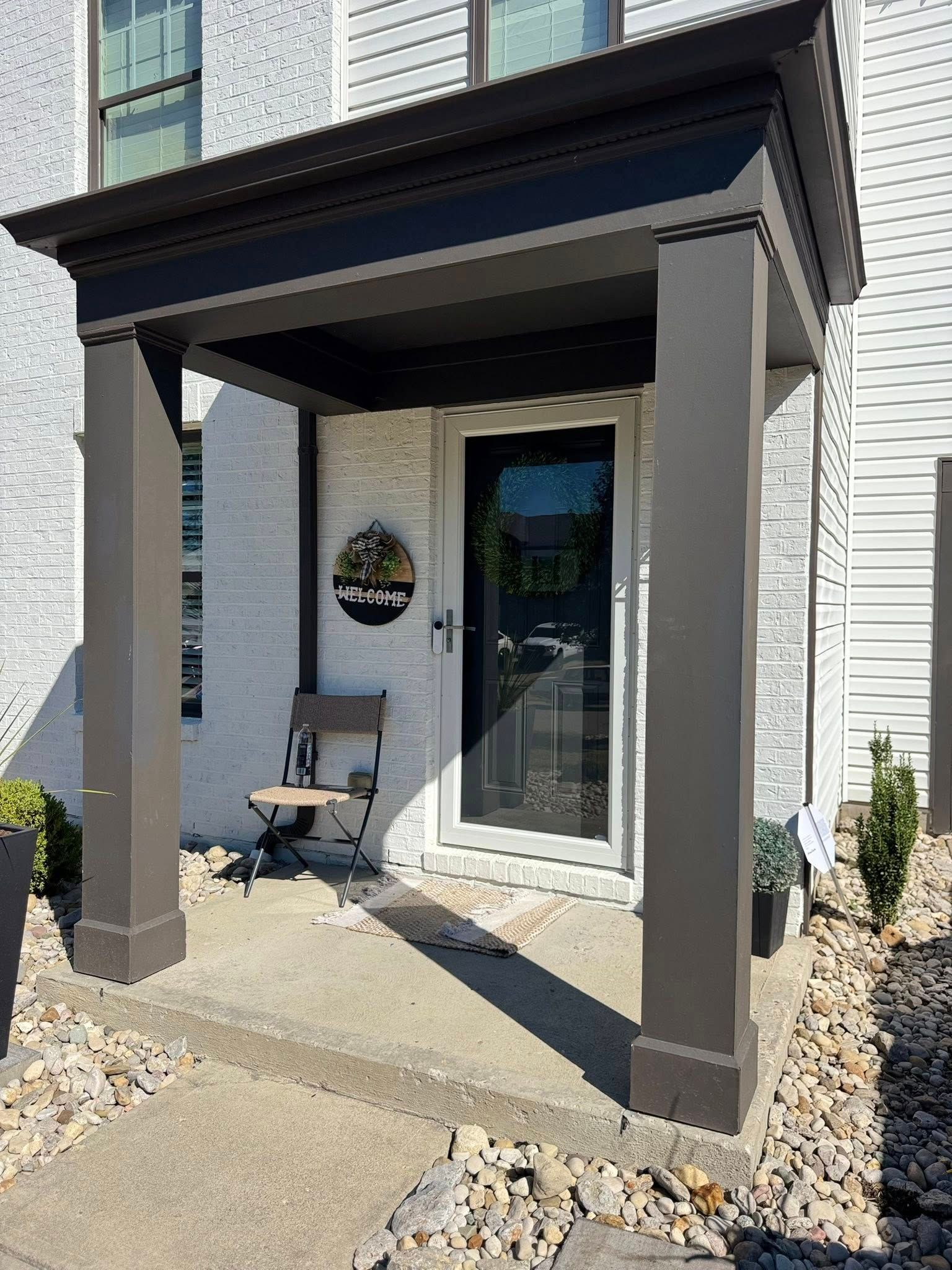 Gray entry porch with dark brown columns and canopy, front door, chair, potted plants.