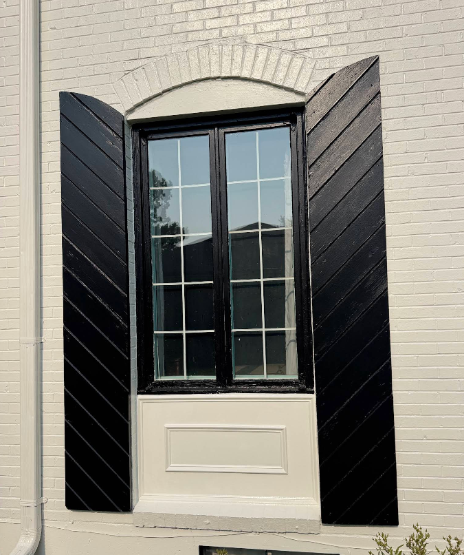 Window with black shutters on a white brick wall. The window has a divided pane and arched top.
