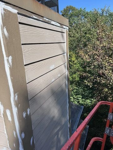 Beige siding on a building's side, next to a green tree backdrop, with a lift in view.