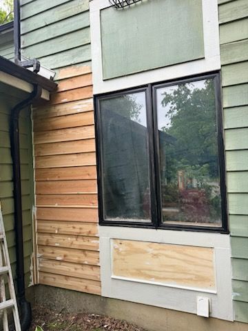 Exterior house wall with two-pane black-framed window; siding is partially unfinished cedar and green.