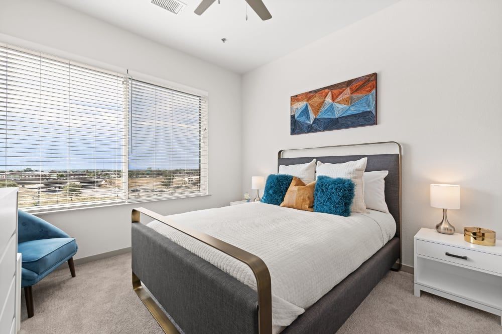 Modern bedroom interior with a neatly made bed featuring a gray frame and white linens, complemented by blue and brown decorative pillows. An abstract geometric wall art hangs above the bed, matching the blue armchair in the corner. The room is brightened by natural light from a large window with closed blinds, showcasing a view of the outdoors. Beside the bed are two white nightstands, each with a simple lamp, set against a well-carpeted floor and minimalist decor at Marq Iliff Station in Aurora, CO.
