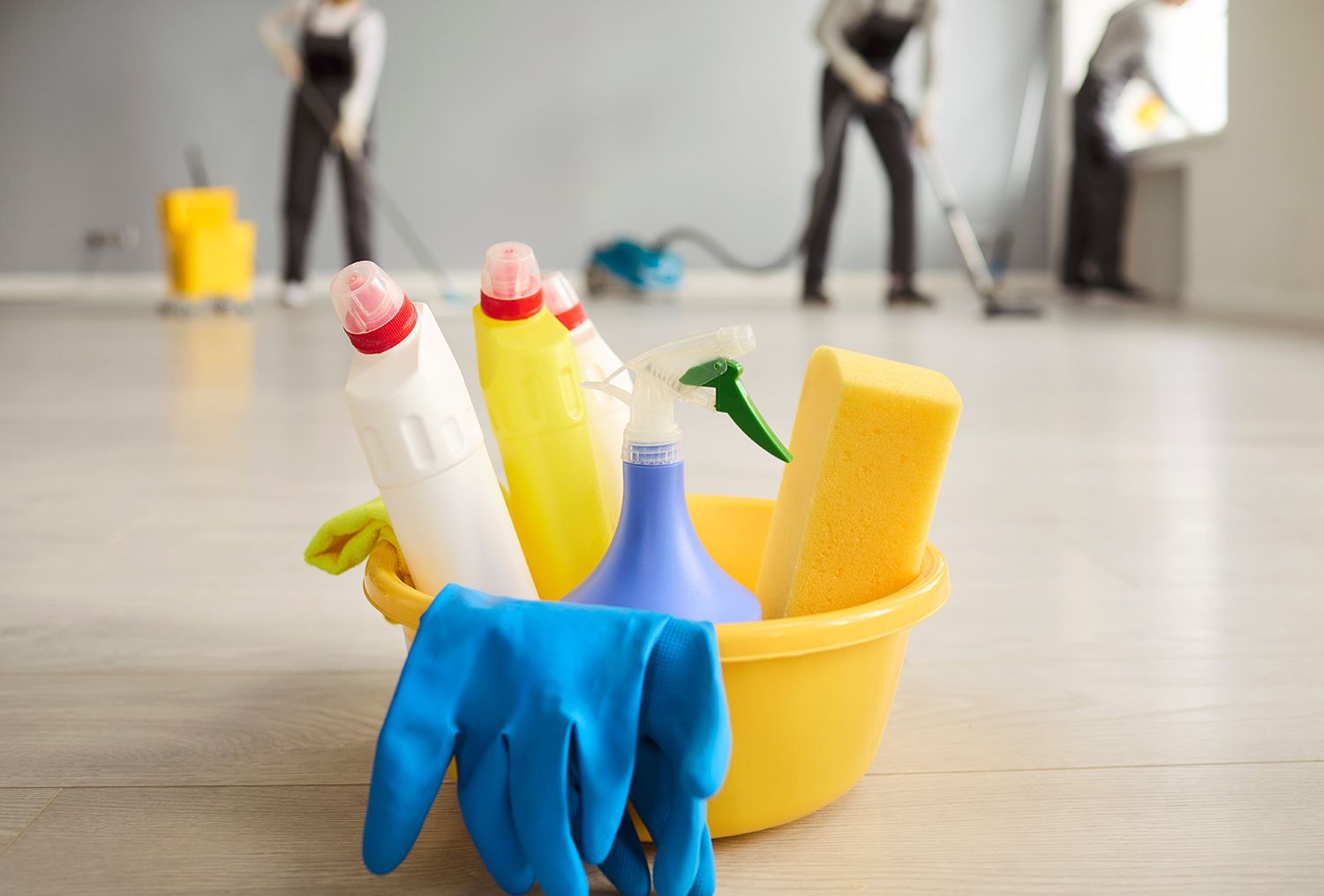 Cleaning supplies in a bucket with blue gloves on a floor, workers blurred in the background Cleaning supplies in a bucket with blue gloves on a floor, workers blurred in the background