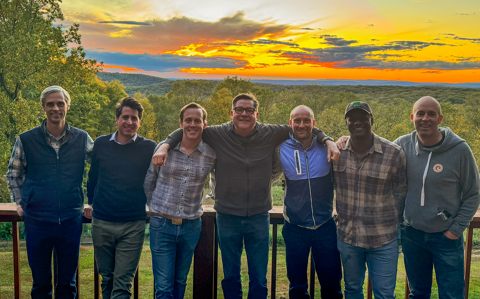 Group of seven Southfield Partners with arms around each other, smiling, against a sunset backdrop over trees.