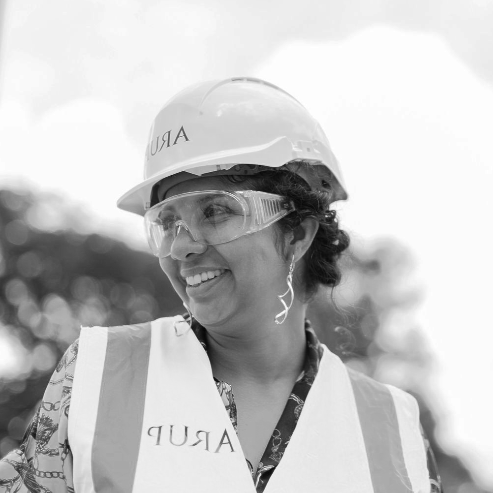 Woman in safety gear smiles, wearing a hard hat, safety glasses, and reflective vest outdoors.