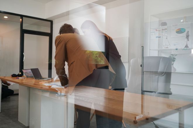 Man in a brown jacket leans over a desk with another person in a bright office setting.