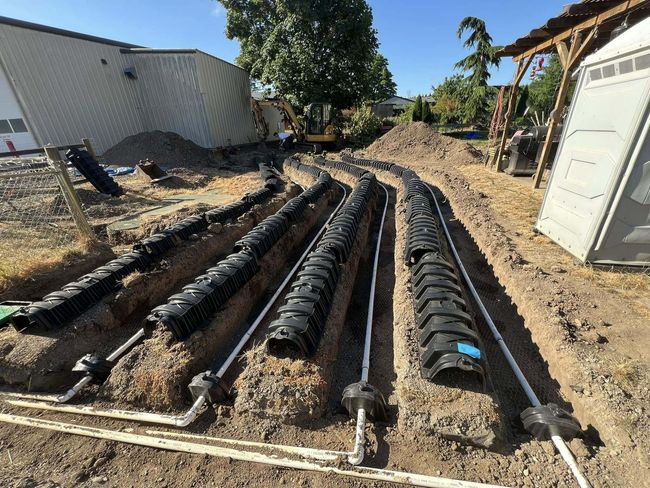 Black drainage pipes laid in a trench beside a house, with dirt and construction equipment nearby.