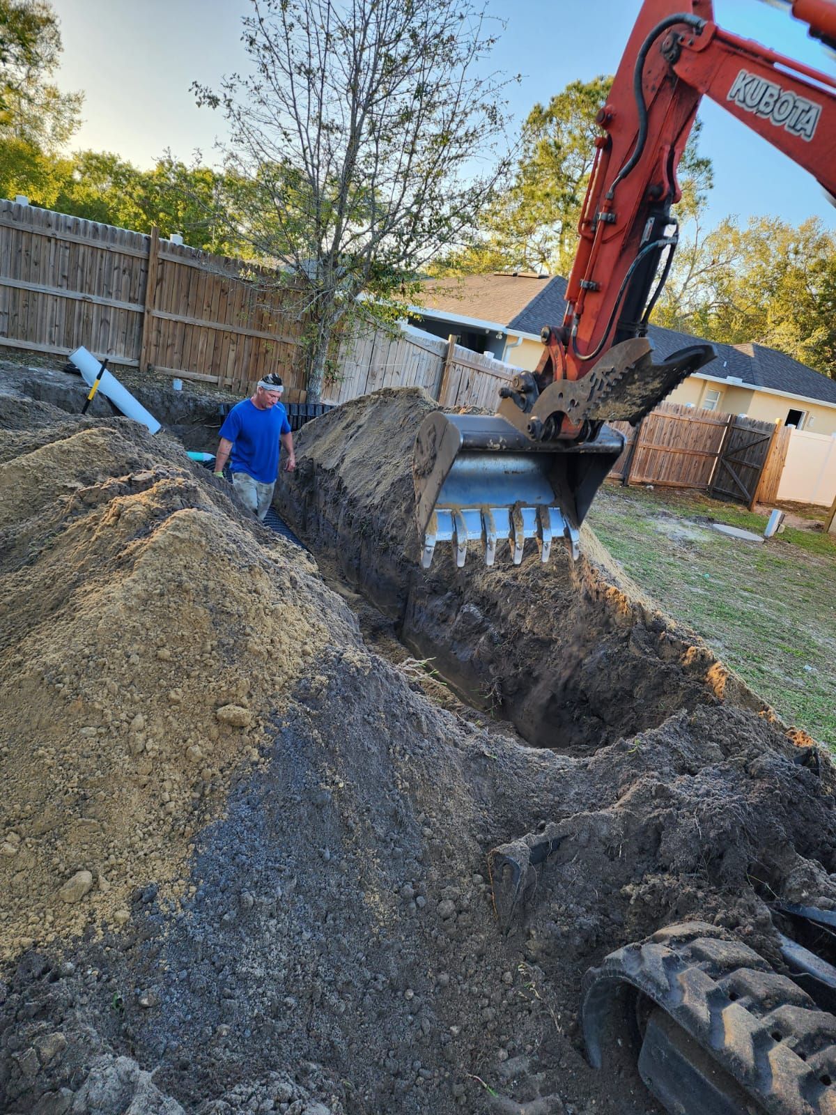 Excavator digging a large trench beside a worker in blue near a fenced backyard at dusk