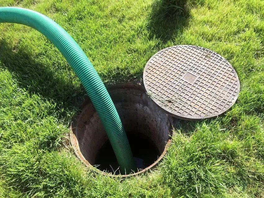 Green hose feeding into an open manhole beside a round metal cover in grass