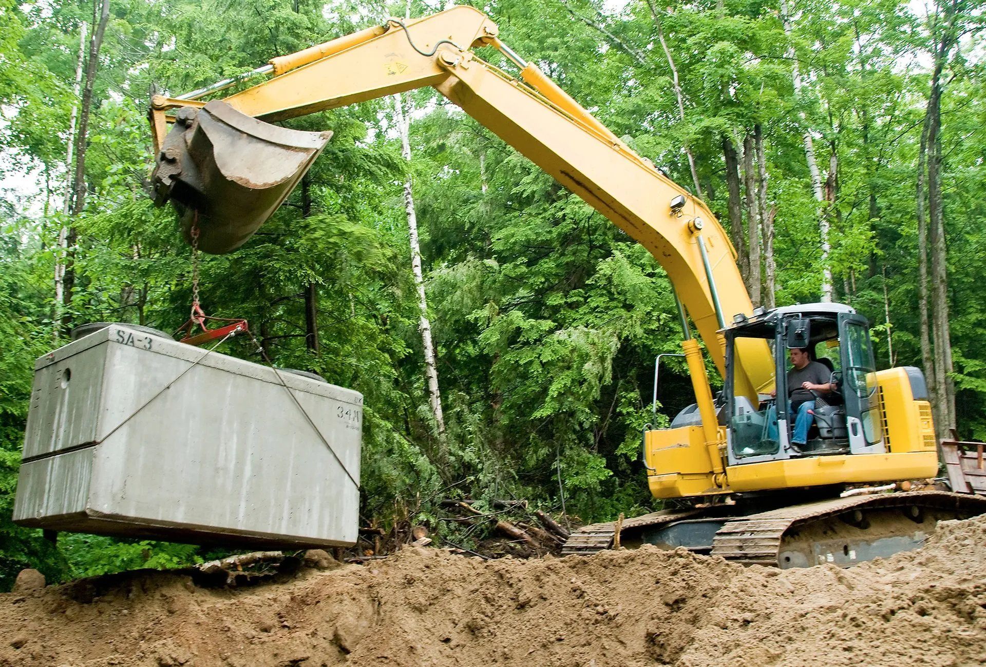 Yellow excavator lifting a concrete block in a wooded construction site