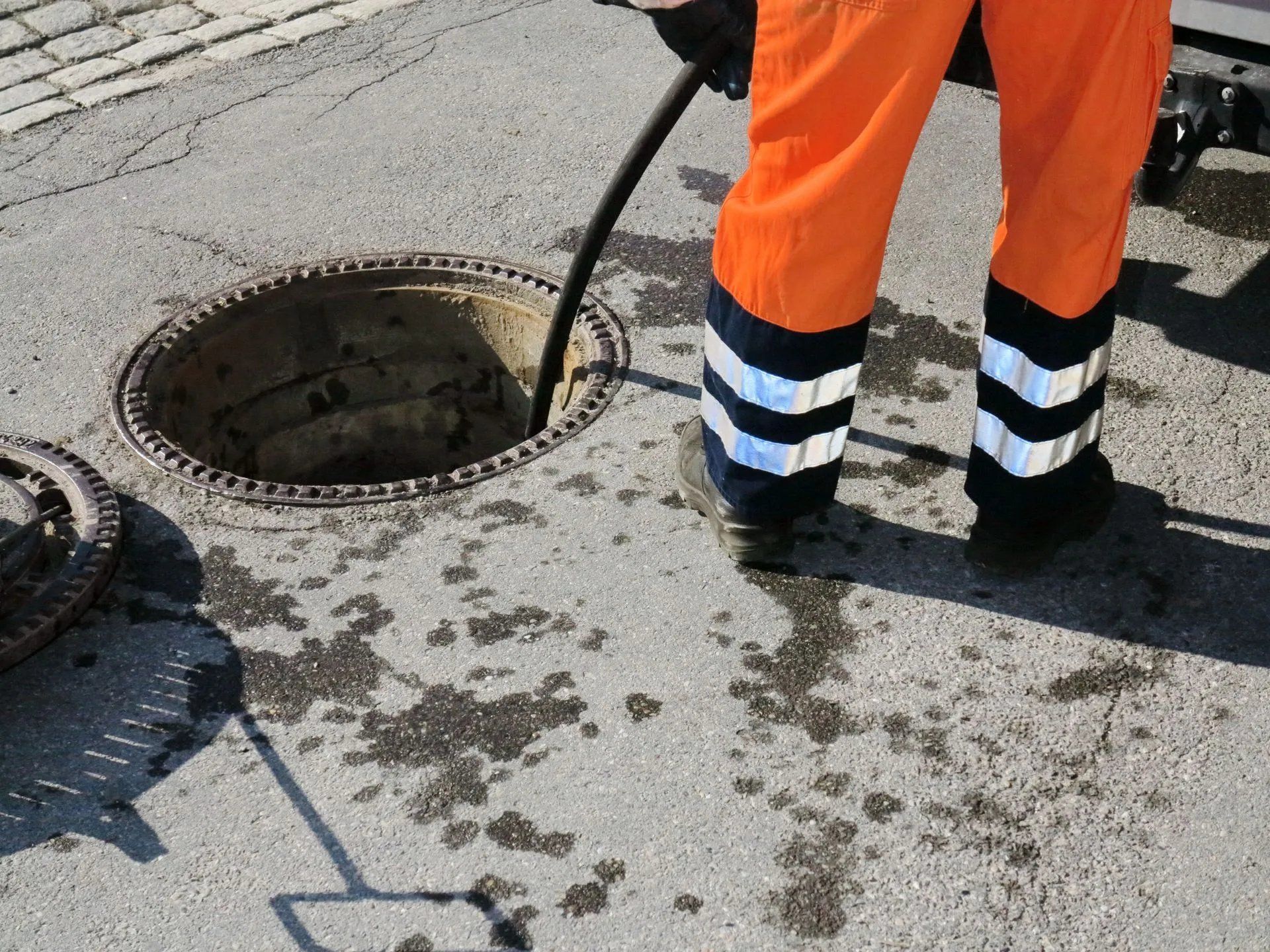 Worker in orange pants stands by an open manhole on a wet street.