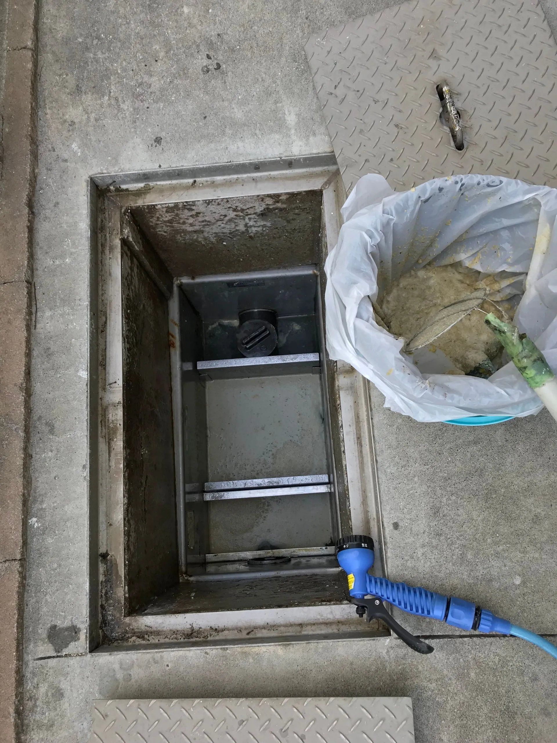 Open utility hatch in concrete sidewalk beside a trash bag and blue tool handle