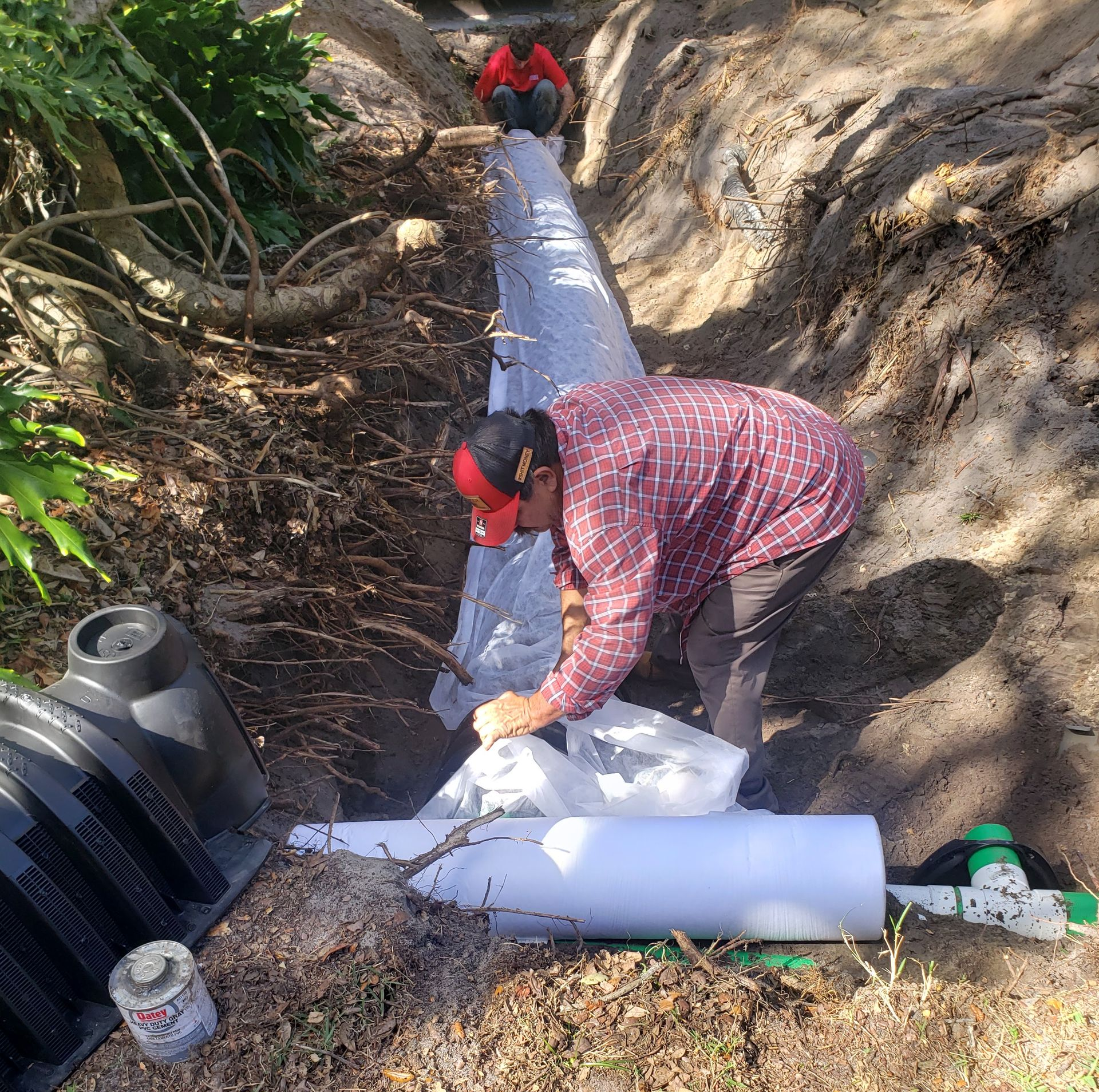 Two workers laying white pipe in a narrow dirt trench on a hillside.