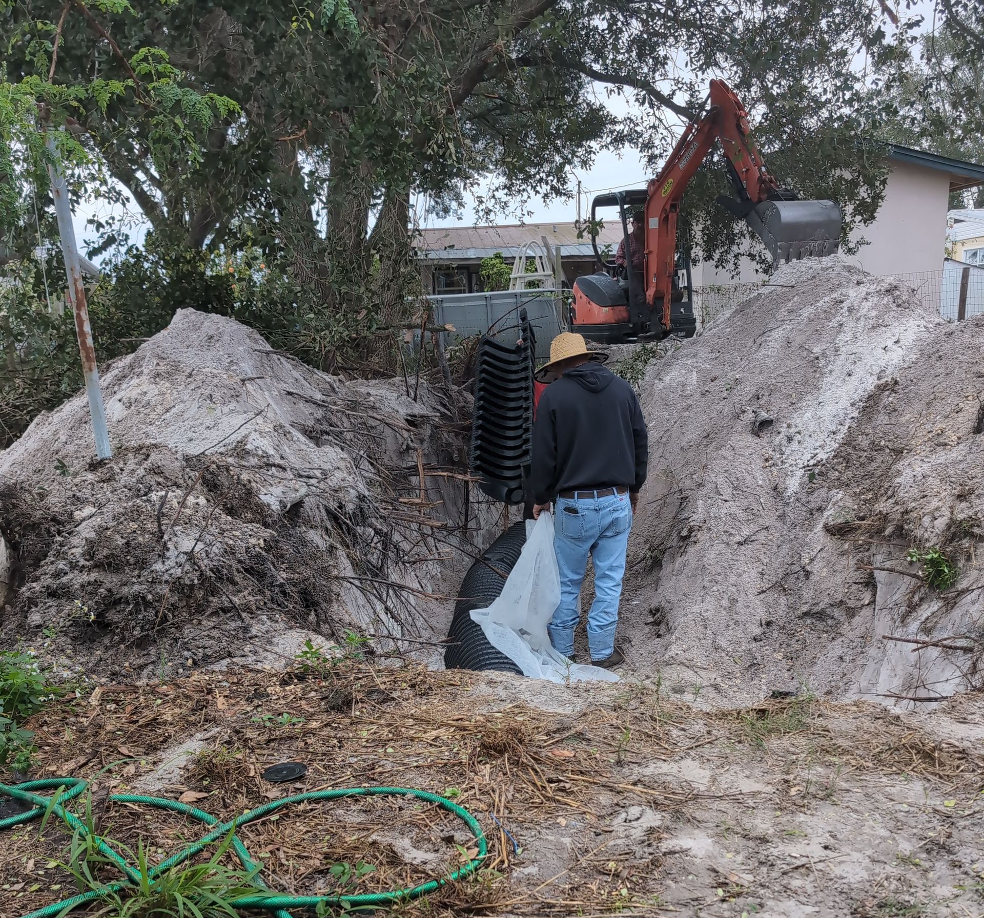 Worker beside excavator in a muddy trench between two dirt piles, with trees and a house in the background