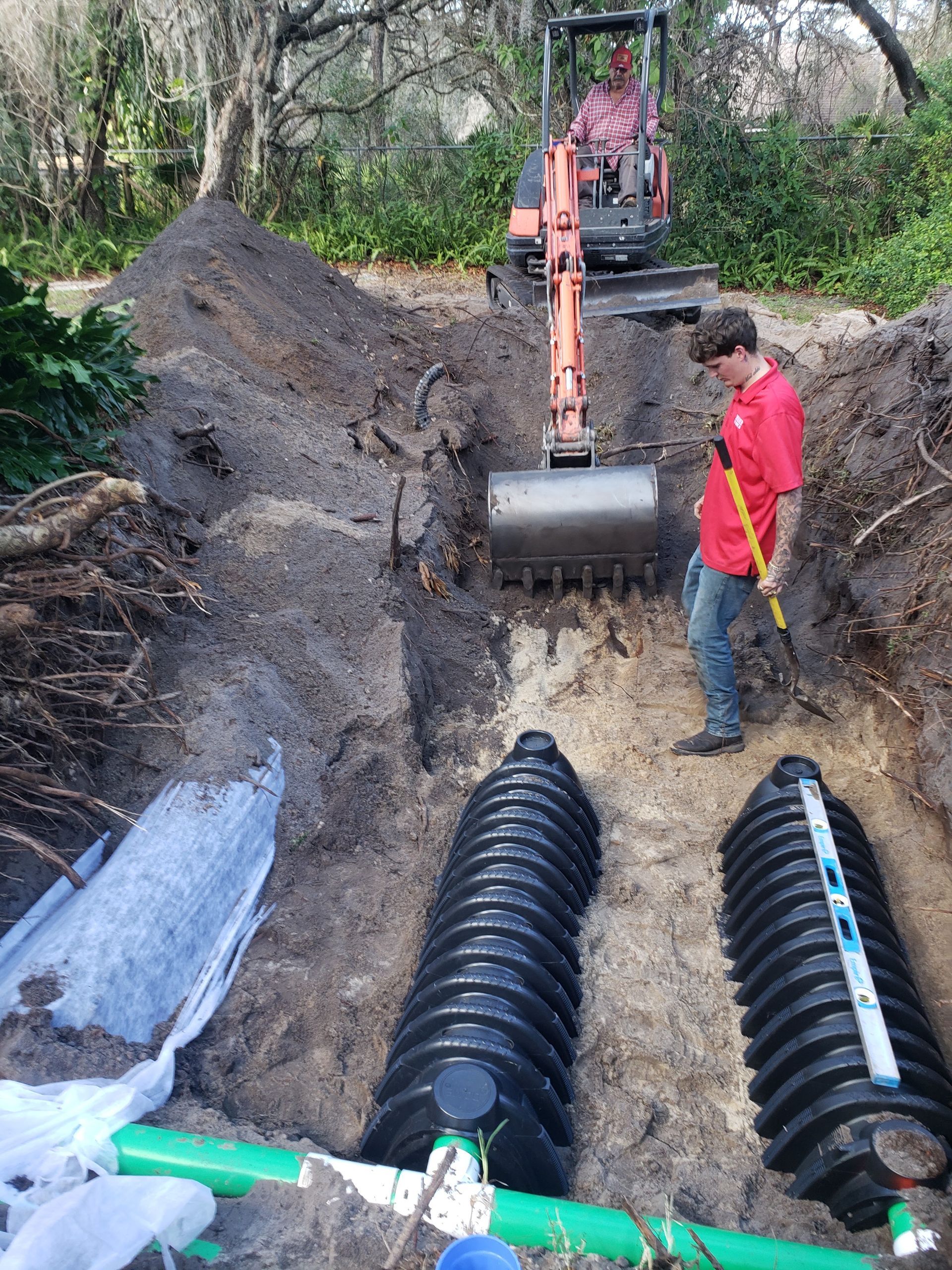 Excavator and worker installing black drainage pipes in a dug trench outdoors