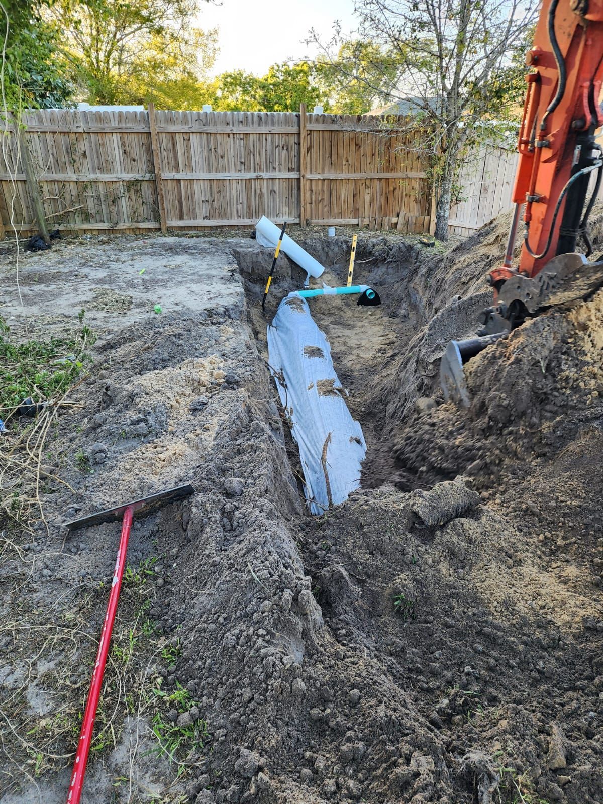 Backyard trench with a gray pipe, dirt piles, and an orange excavator beside a wooden fence.