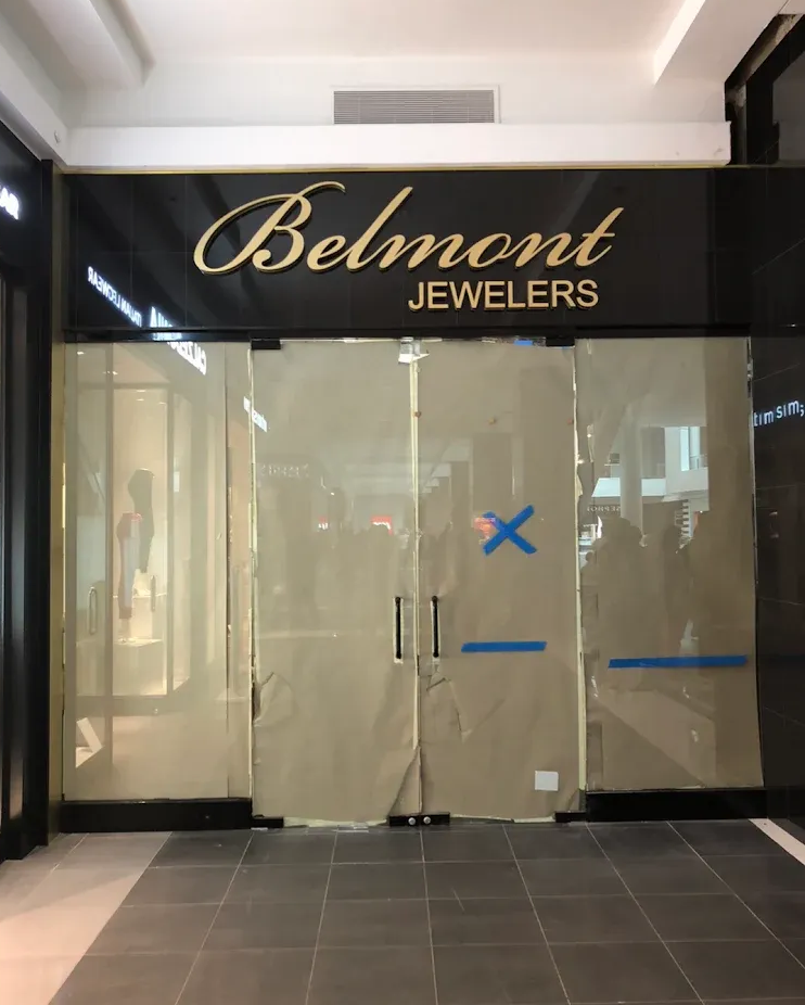Closed Belmont Jewelers store front, gold lettering on black, windows covered, blue tape and X.