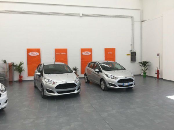 Autosalone con due auto argentate su un pavimento grigio e insegne arancioni sulla parete di fondo.