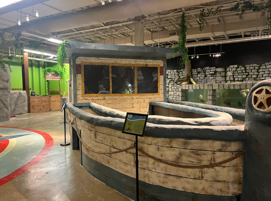 Indoor play area with a rustic boat-themed exhibit, stone wall backdrop, and hanging green vines