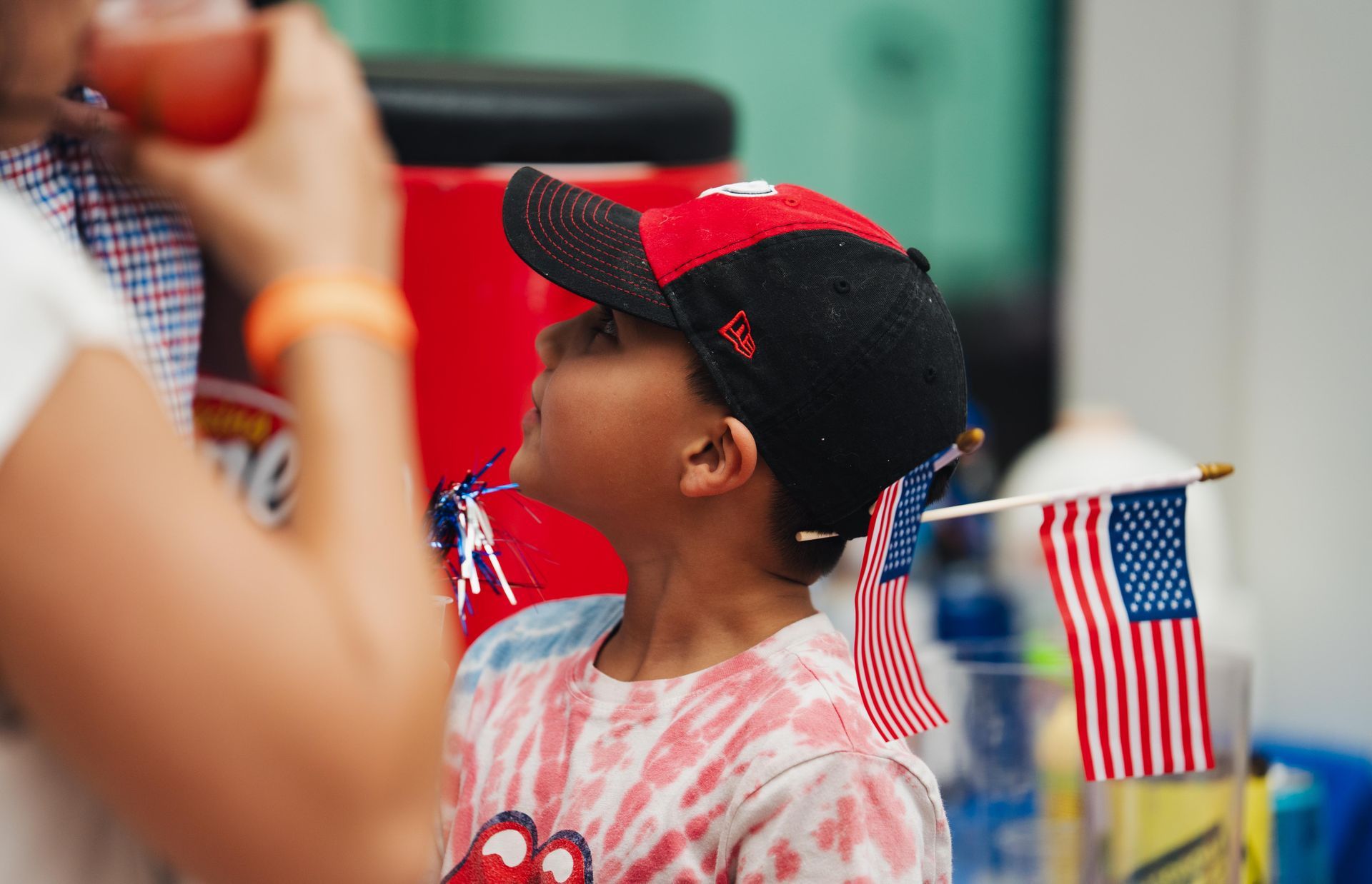 Child wearing a red, white, and blue cap watches a patriotic parade with small American flags nearby