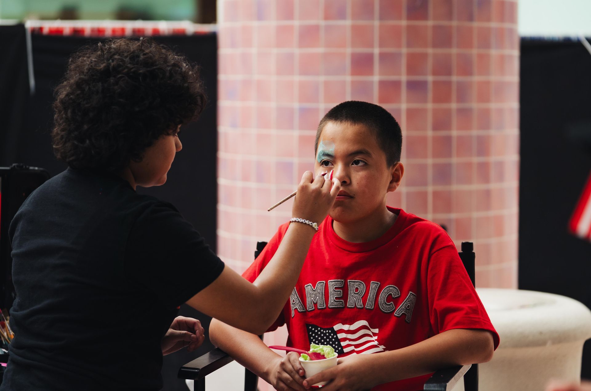 Child in red shirt sits while another person applies face paint outdoors.