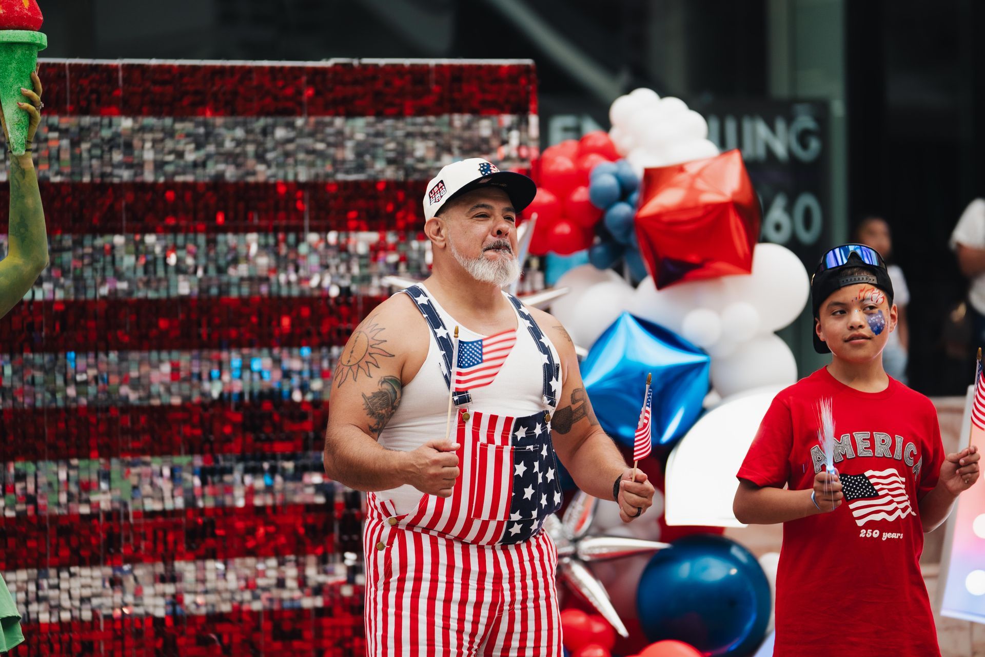 Two people posing outdoors with red, white, and blue balloons and an American flag backdrop