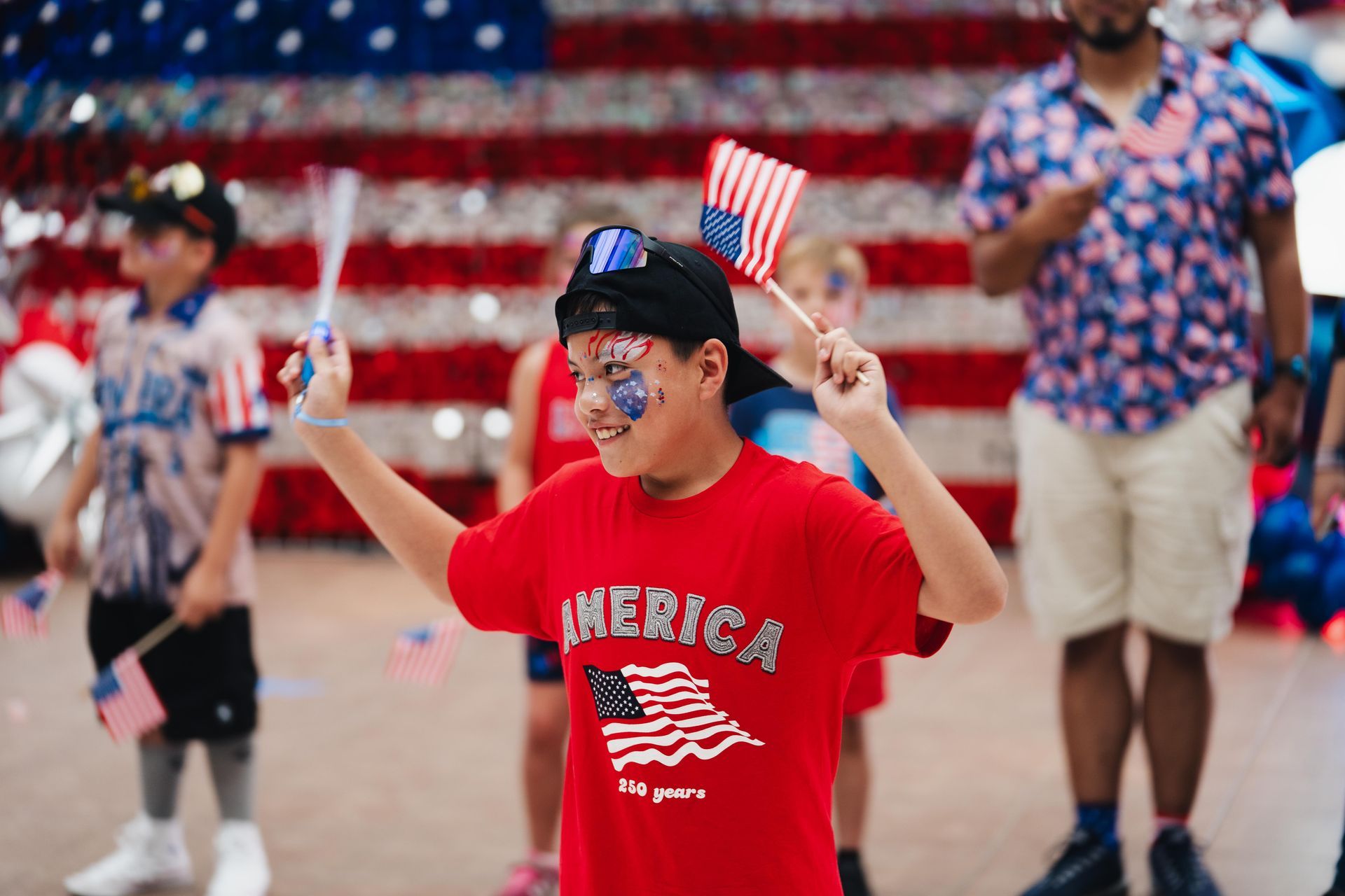 Boy in red “America” shirt holds small flags in a patriotic parade scene.