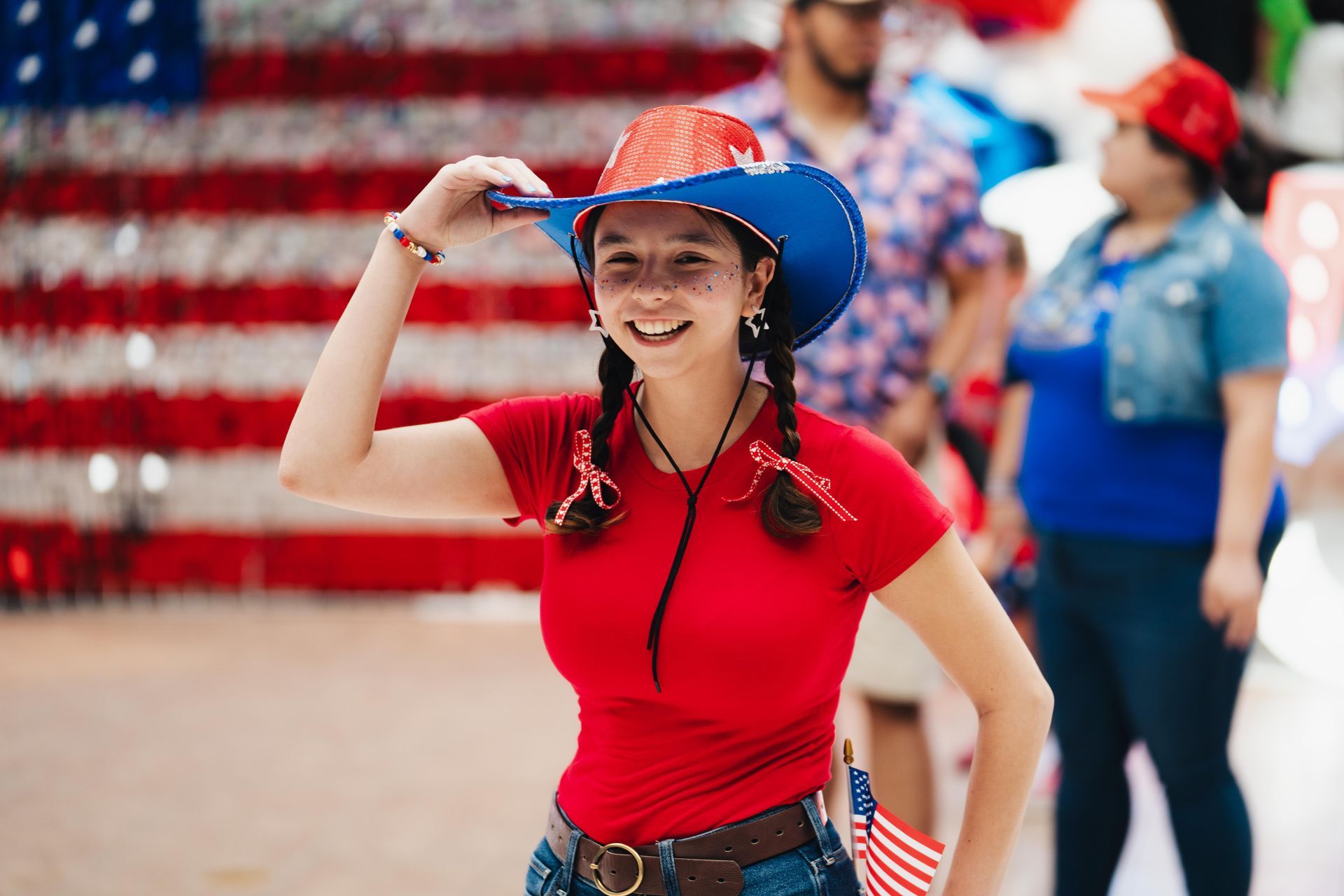 Woman in a red shirt and blue hat saluting in front of an American flag backdrop