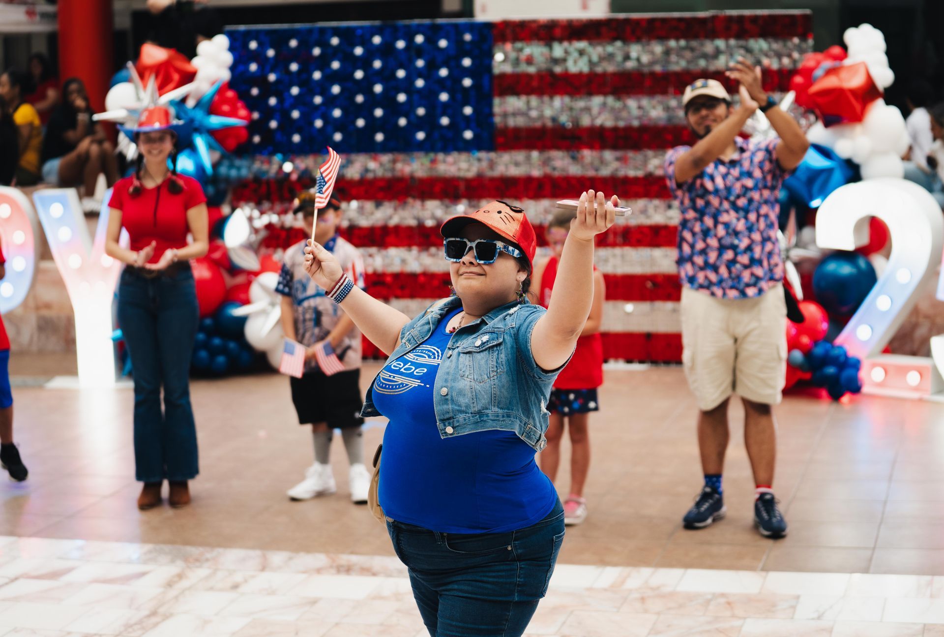 Dancers in red, white, and blue costumes perform in a patriotic indoor celebration with a U.S. flag backdrop