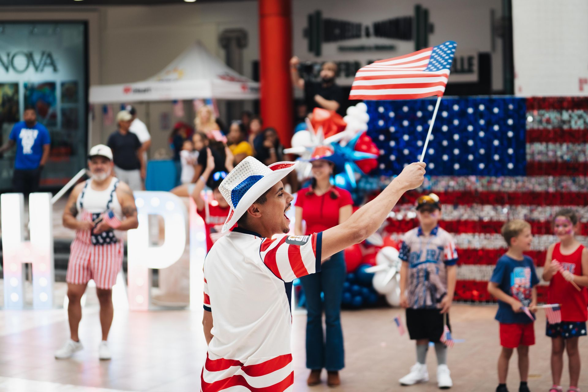 Costumed performer waving a small American flag in a lobby, with children and a large U.S. flag behind him.