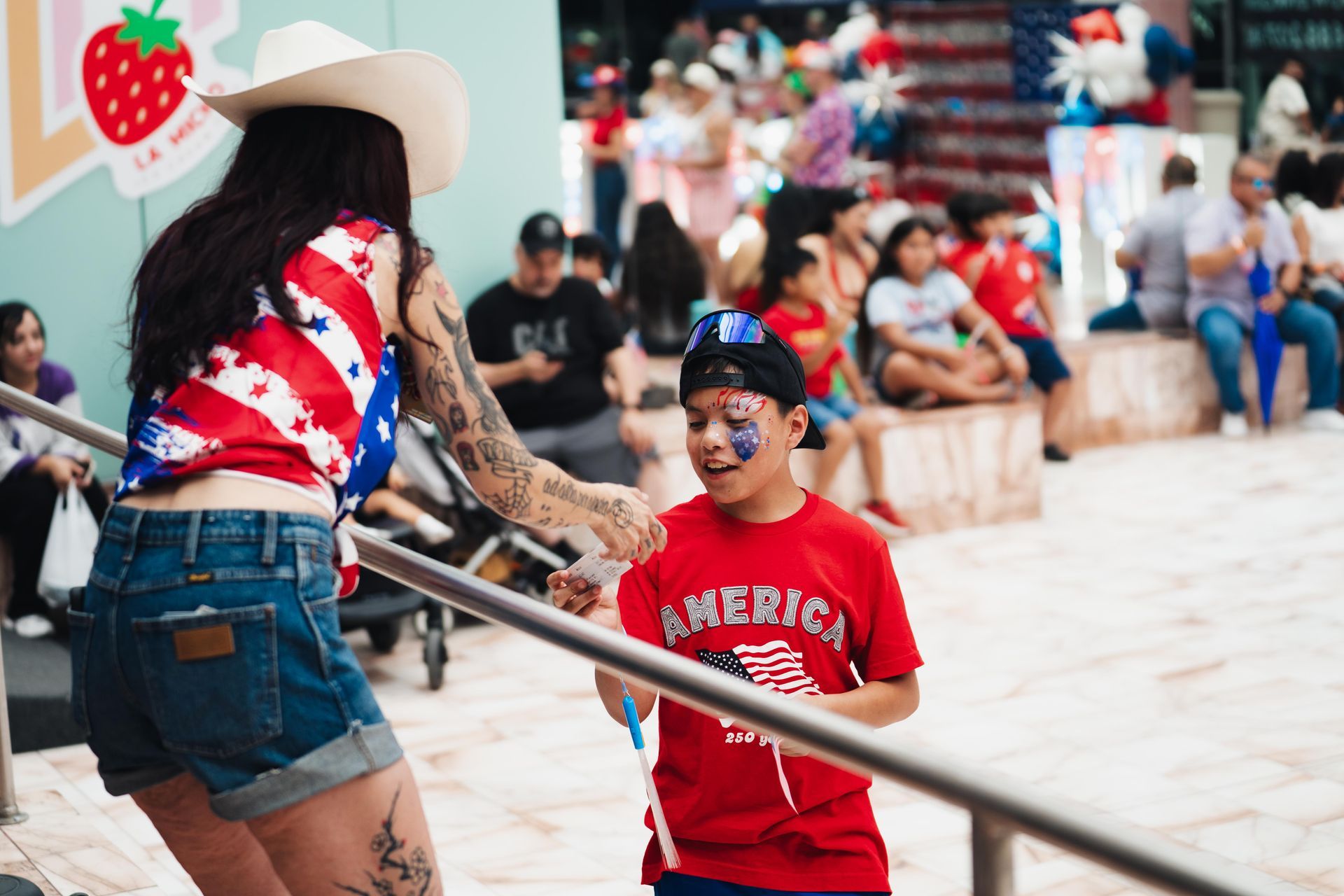 Woman in cowboy hat and child in red shirt at a busy outdoor event, standing by a metal railing