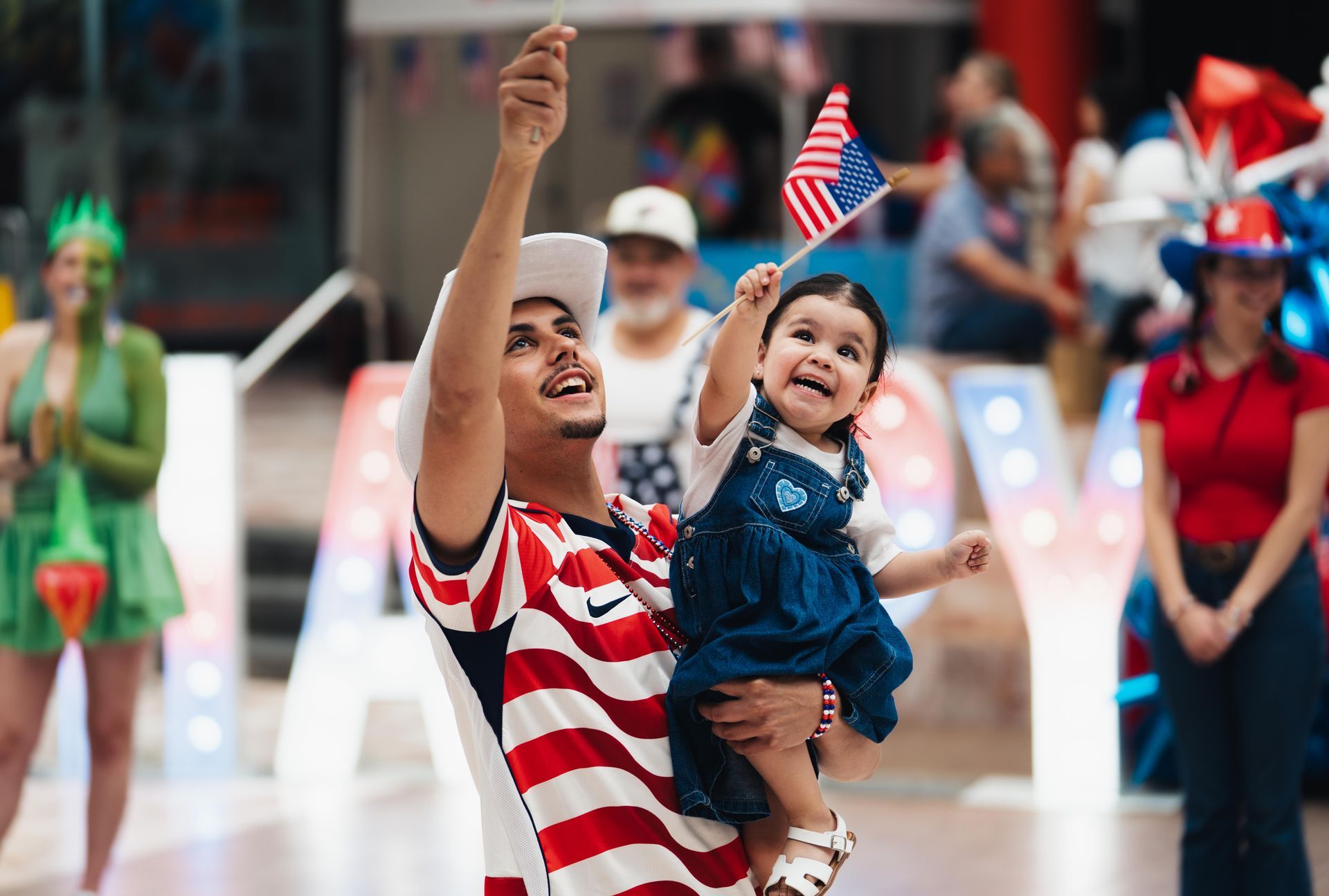 Man holding a waving child in a denim dress, both smiling amid a festive crowd and flags