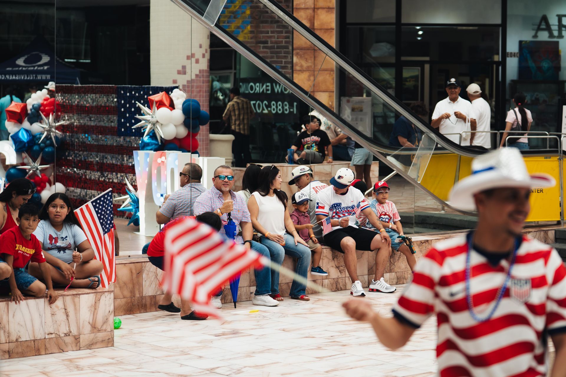 People sitting on a sandy beach waving American flags near a building with red, white, and blue decorations