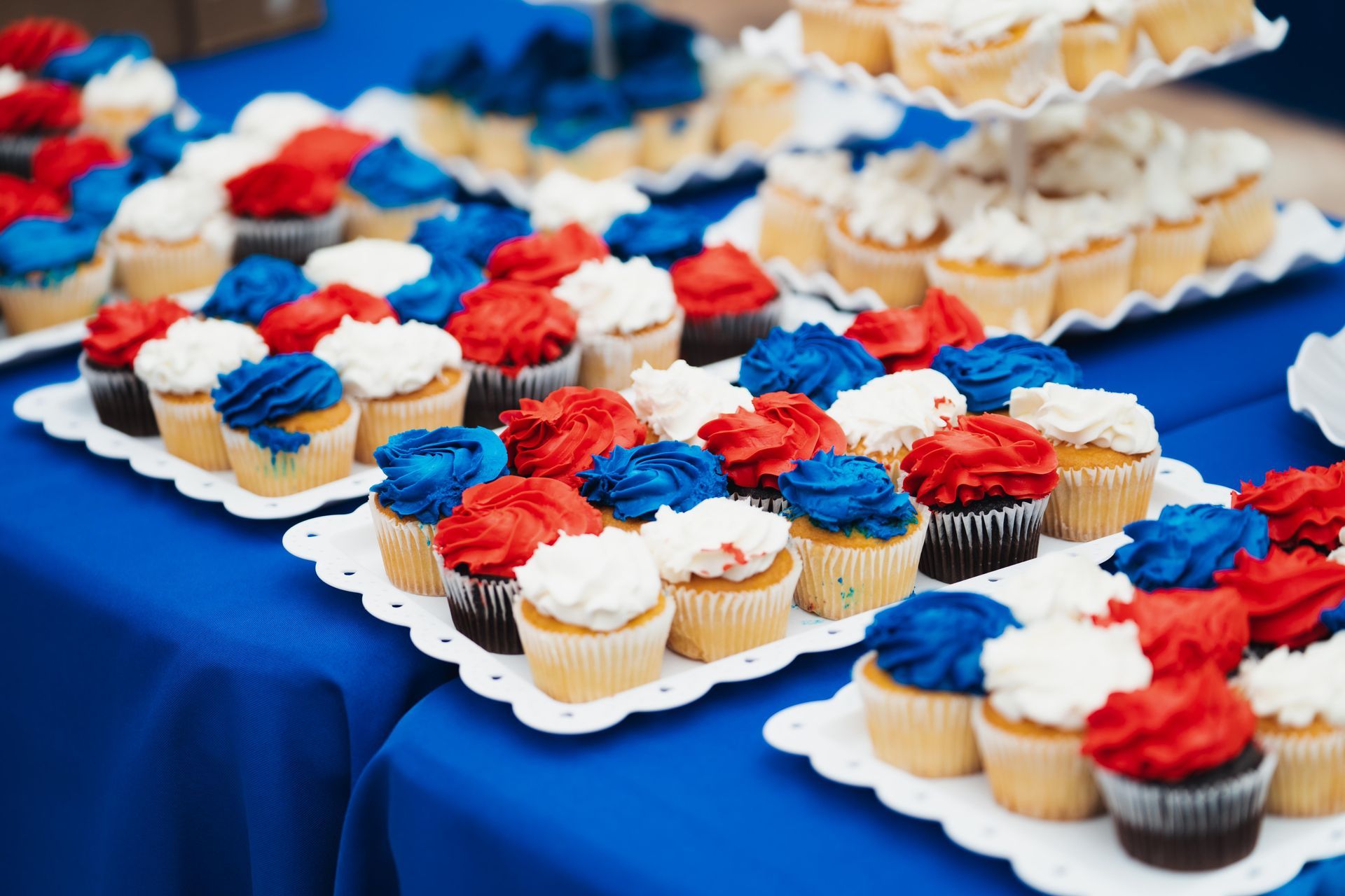 Rows of red, white, and blue frosted cupcakes on trays at a table with a blue cloth
