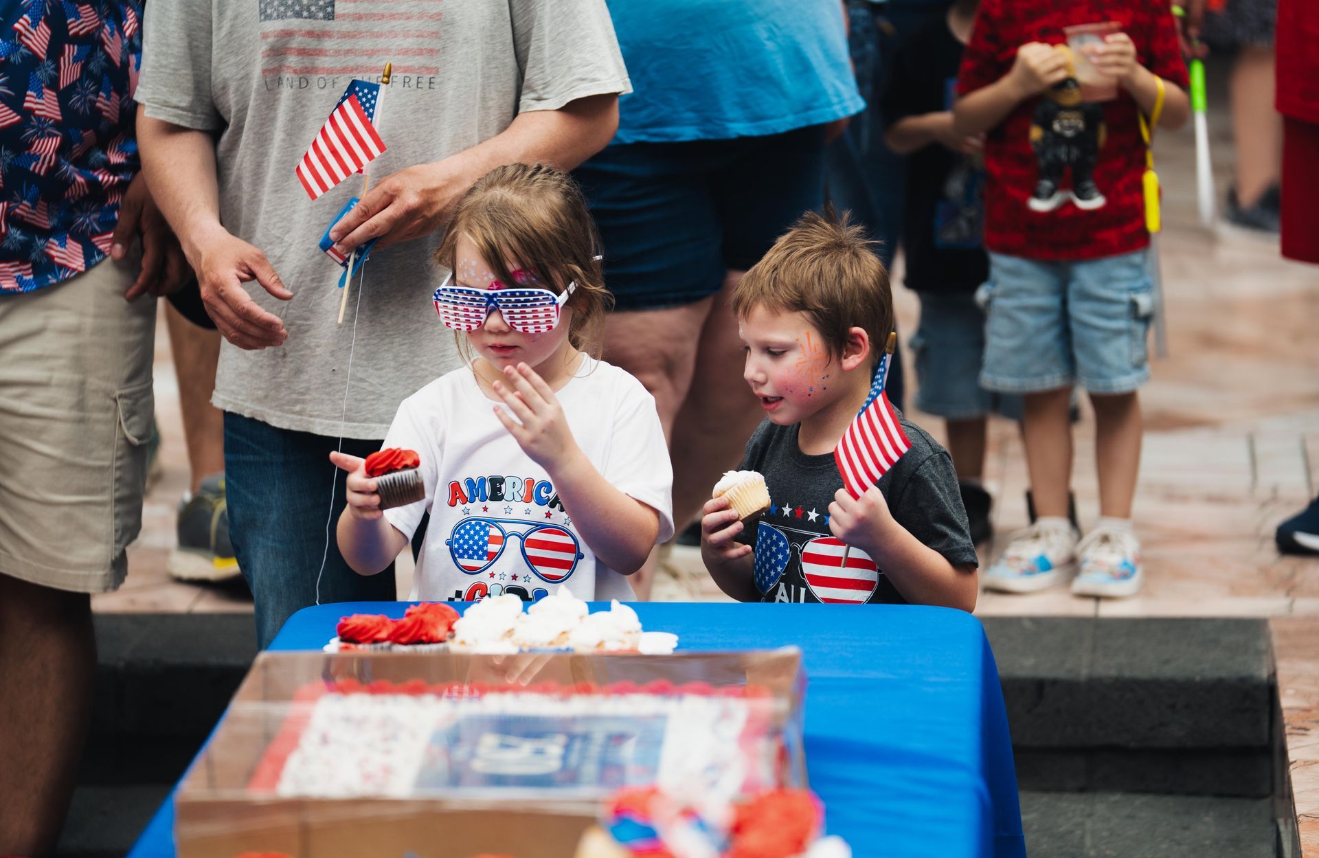 Two children in sunglasses sit at a cake-covered table, waving small flags at an outdoor celebration.