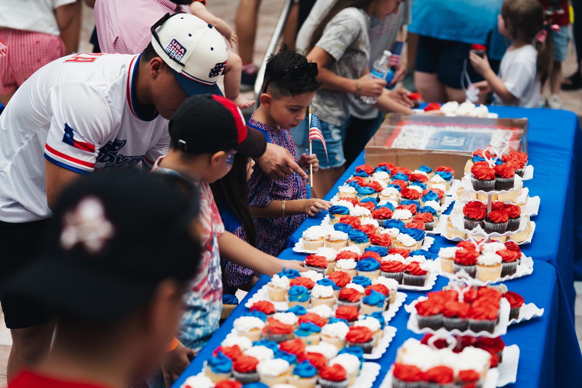 People leaning over a long blue table displaying rows of red-and-white frosted cupcakes at an outdoor event