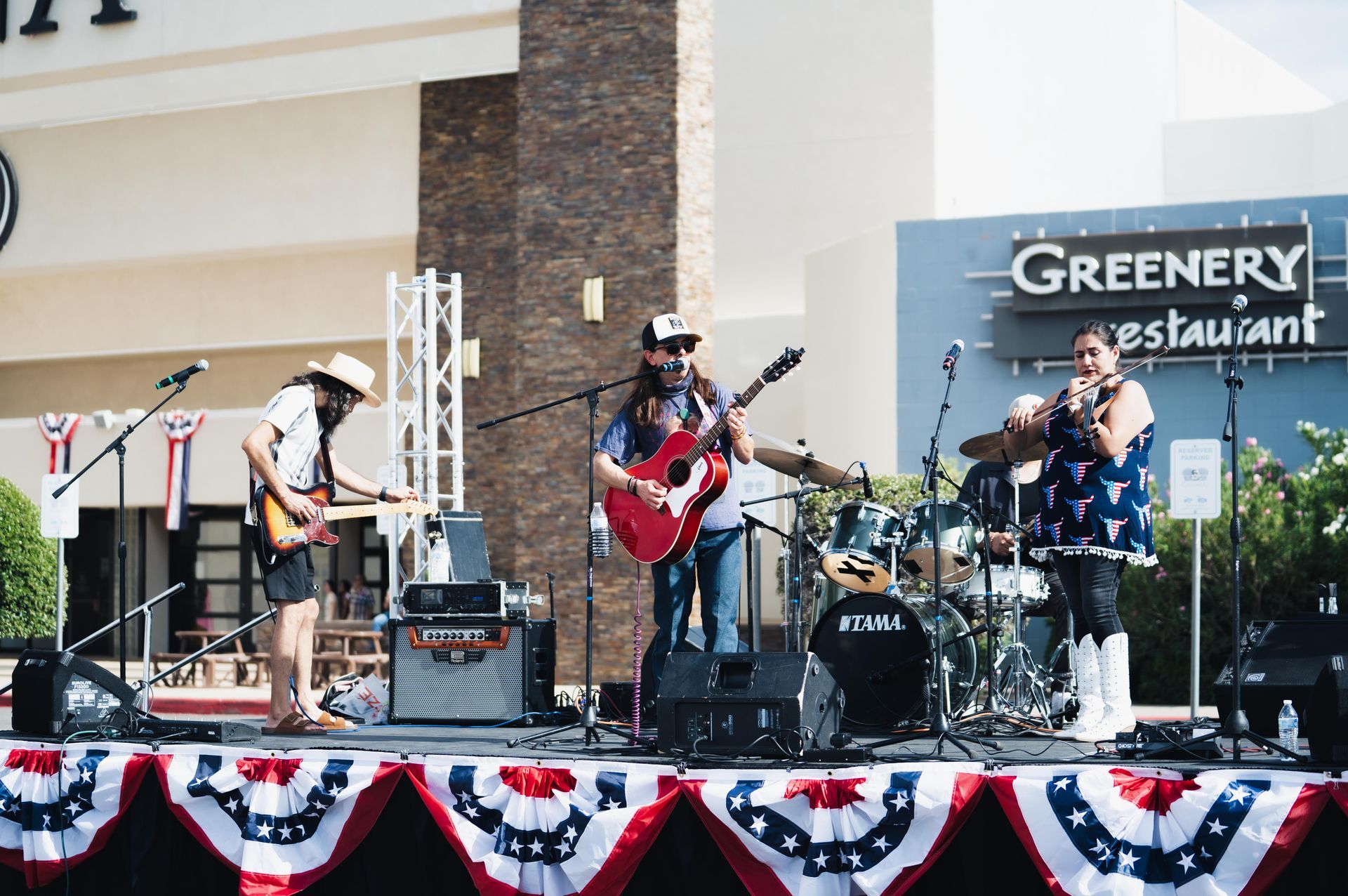 Band performing on an outdoor stage in front of a Greenery restaurant sign, with red-white-blue bunting.