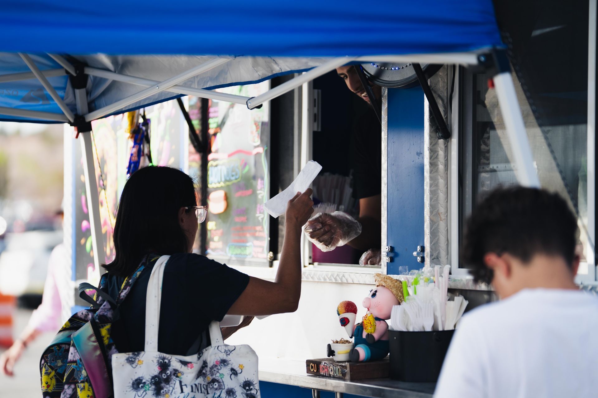 Person at a market stall under a blue canopy handing cash to a vendor at a counter