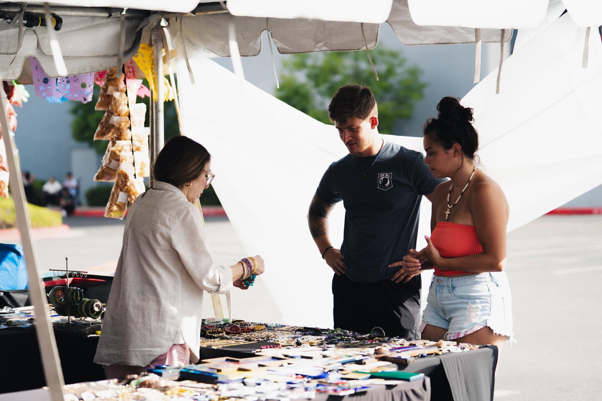 Three people browsing items at an outdoor market stall under a white canopy