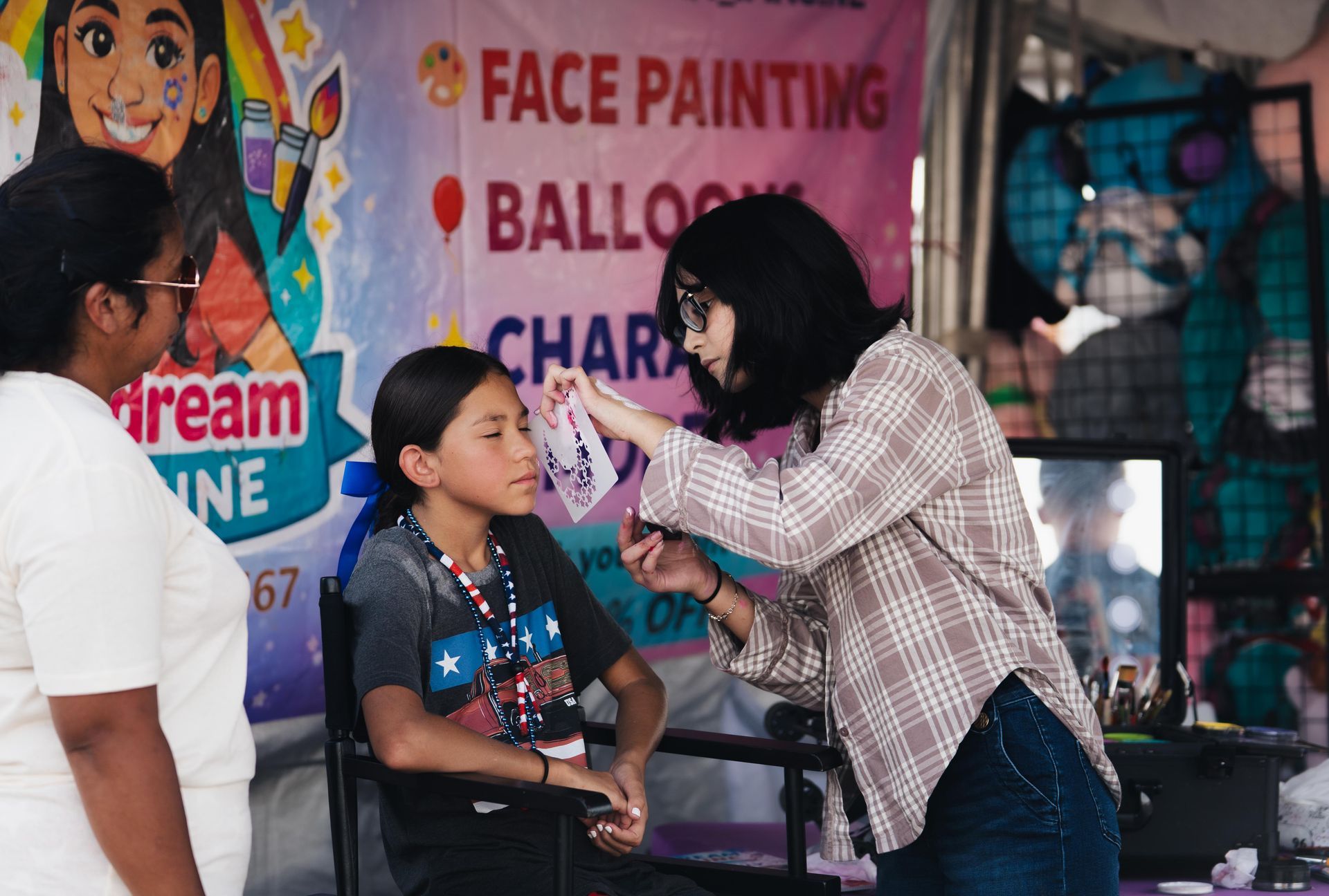 Face painter applying makeup to a seated child at an outdoor event booth.
