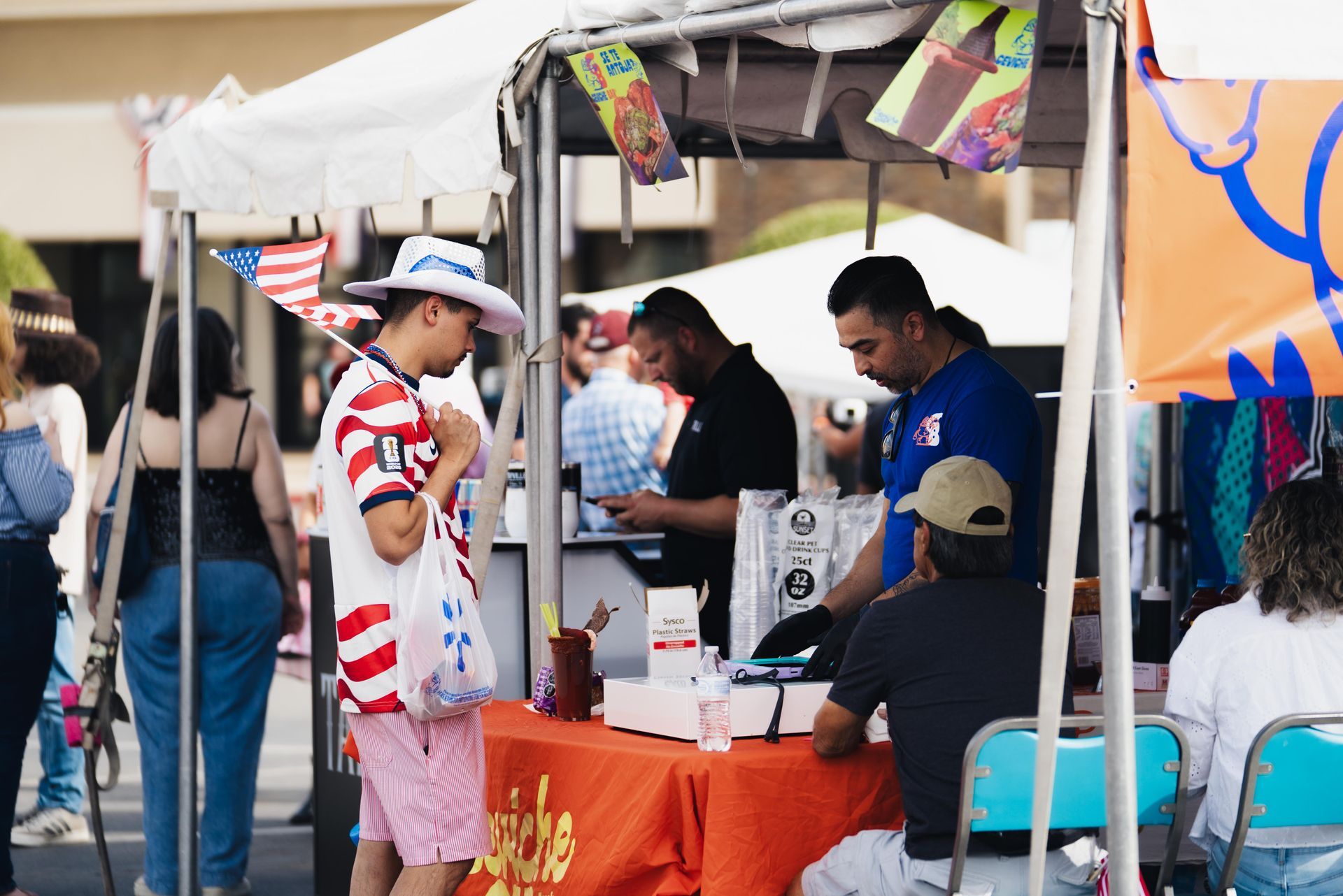 People chatting at a market booth under a white tent, with an orange tablecloth and colorful decorations.