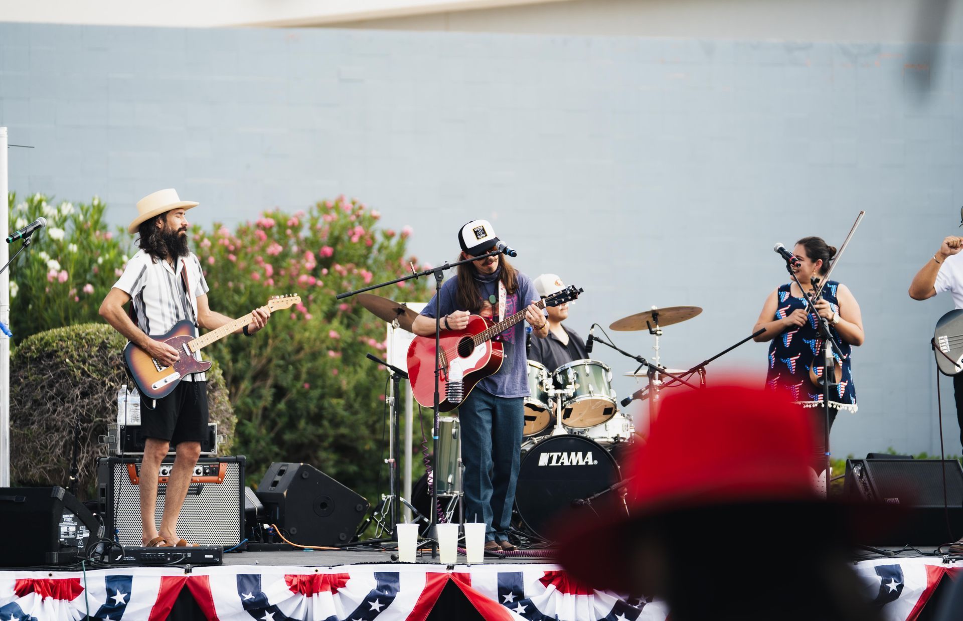 Band performing outdoors on a stage, with guitars, drums, and microphones in front of a plain backdrop.