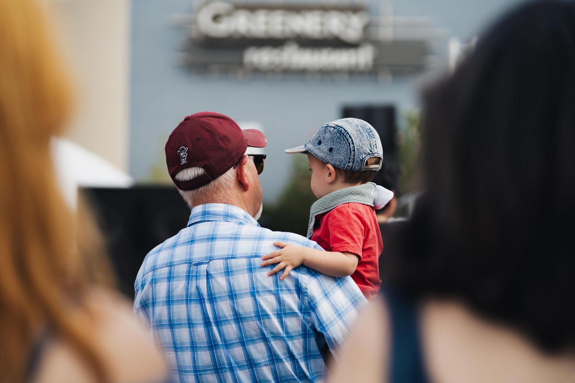 Adult holding a small child in a cap, viewed from behind at an outdoor event with a blurred sign in the background