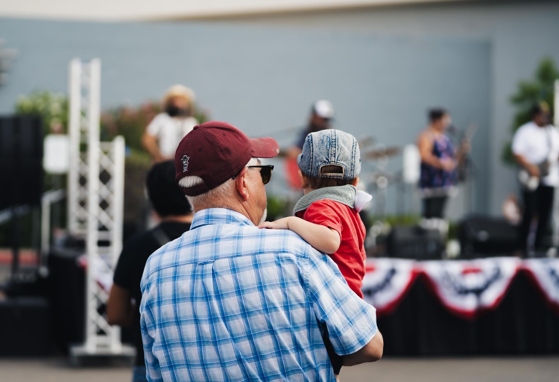 Man holding a child on his shoulders at an outdoor stage event, with people blurred in the background.
