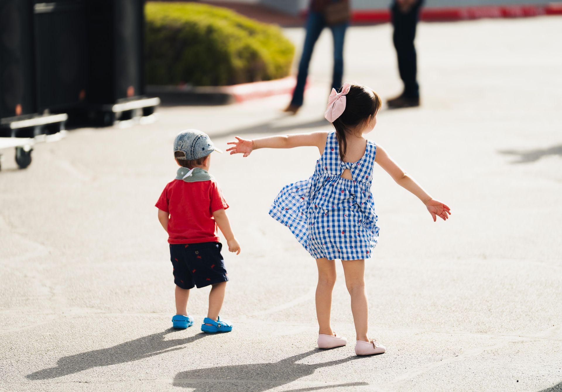 Two children playing outdoors on a sunny street, one in a blue gingham dress with arms outstretched.