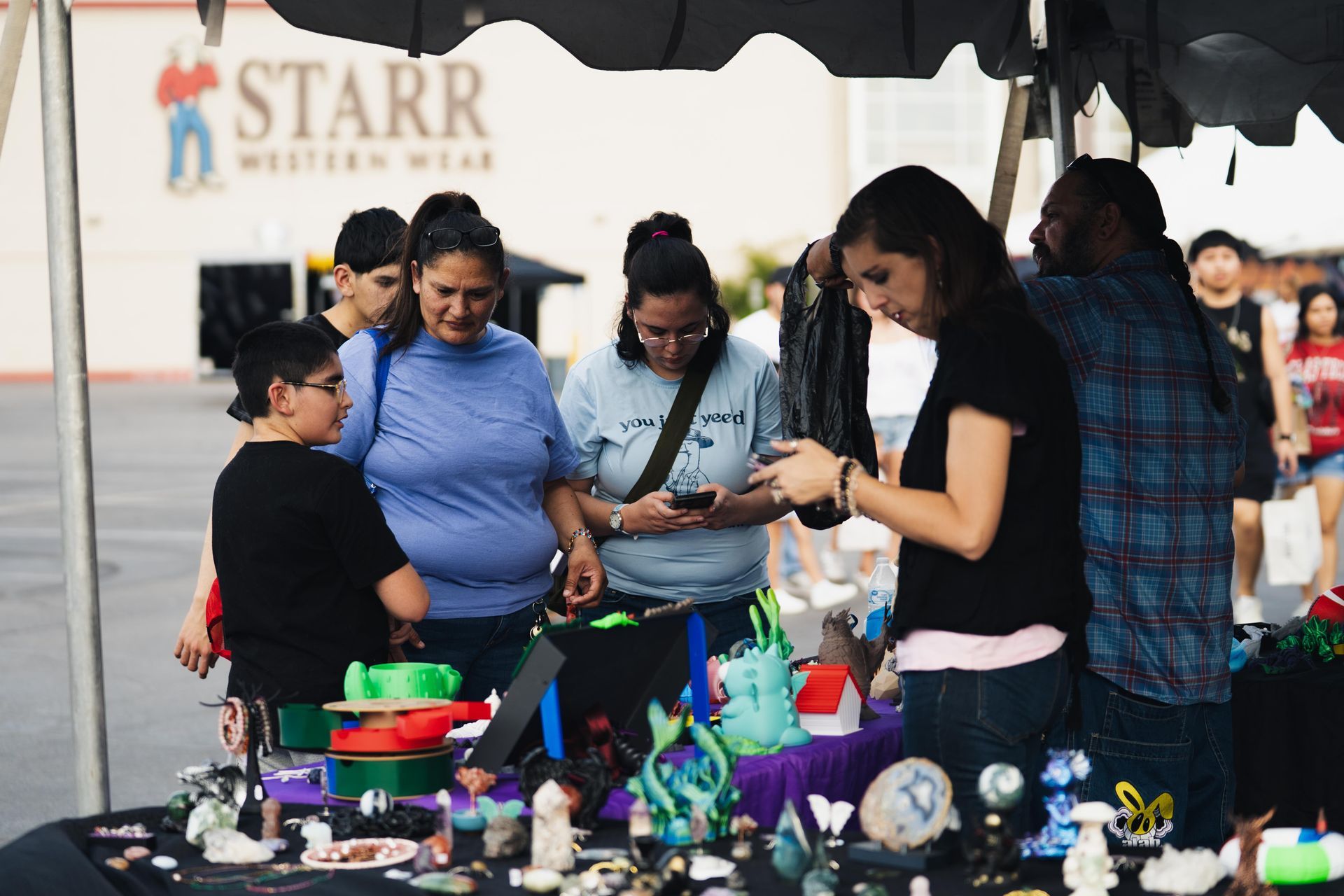 People browsing colorful handmade items at an outdoor market booth under a white canopy