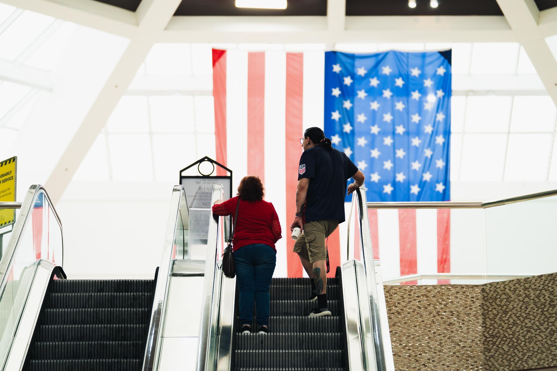 Two people ride up an escalator before a large hanging American flag indoors.